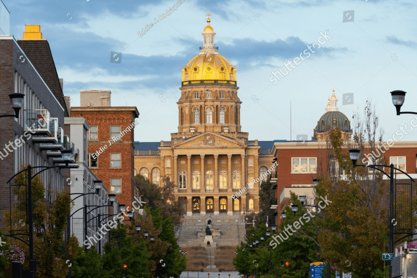 The Iowa State Capitol building with a gold dome is viewed from a street lined with trees and modern buildings in Des Moines, Iowa.